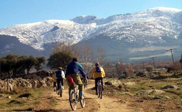 Unos ciclistas pasean a los pies de la sierra de Guadarrama y con el pico de la Mujer Muerta al fondo.